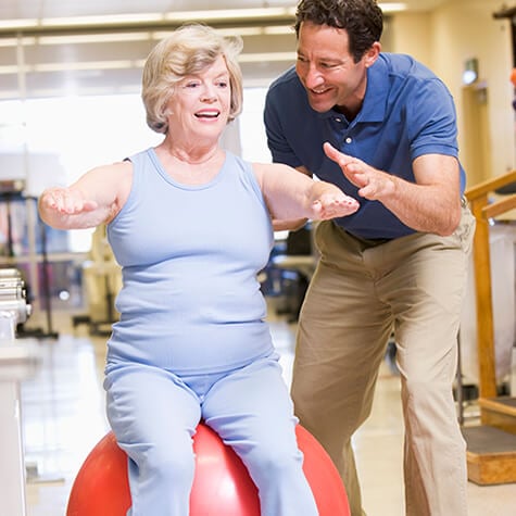 Therapist spotting an older female sitting on a large balance ball with her arms stretched in front of her