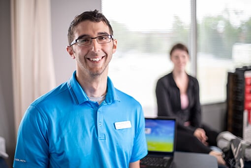 Male therapist wearing a teal blue shirt and name tag standing in front of a laptop on a desk.