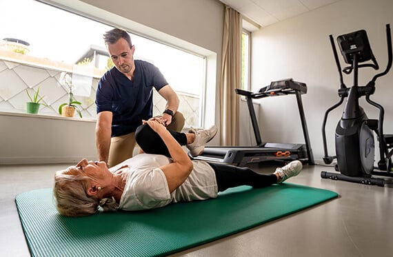 Patient laying on her back on the floor while a therapist helps her bring one knee to her chest